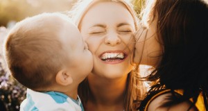 Close up portrait of lovely young mother laughing with closed eyes while her kids is kissing her on the cheeks outdoor against sunset.